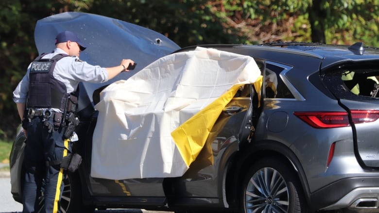 A police officer holds up a phone to a car with a yellow tarp on it, with the car having visible damage.