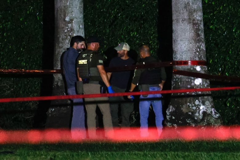 A group of law enforcement officials stand behind police tape in a treed area at night.