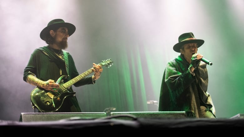 Two musicians, both wearing hats, are surrounded by bright lights while performing on stage.