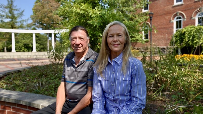 Two people sit together on a bench outside, in front of a brick building and some shrubbery.