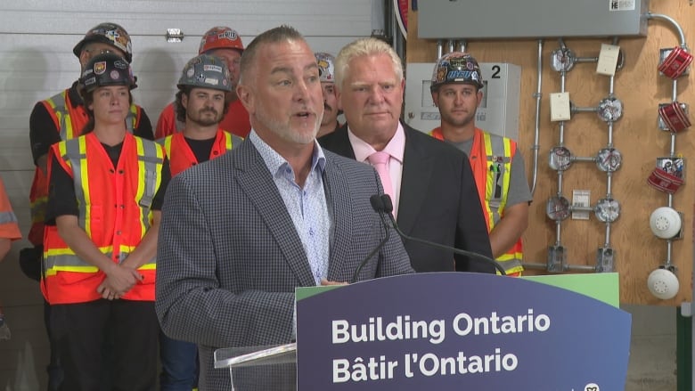 A man in a suit at a podium while workers in hi-viz vests and hard hats stand behind him.