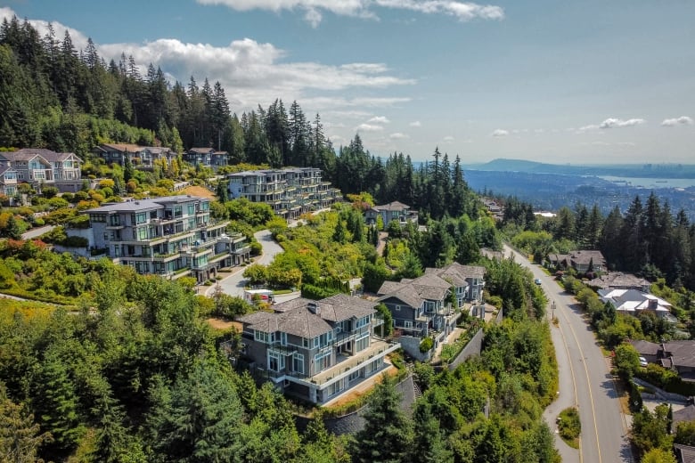 Numerous small apartment buildings are pictured on a picturesque hillside, with a water body and mountains visible in the background.