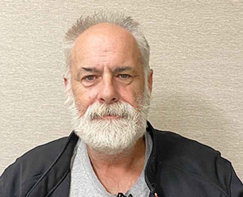 A man with white hair, beard and moustache wearing a dark jacket and grey shirt looks at the camera.