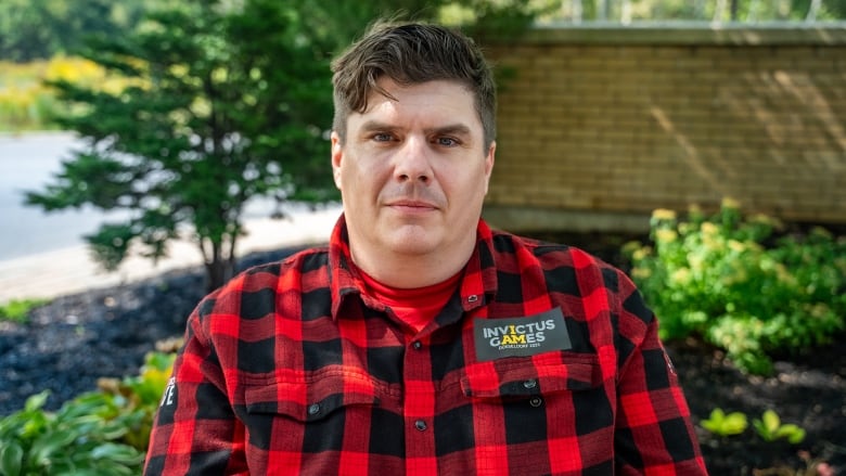 A man in a black and red flannel occupies the frame, the Invictus Games badge over his left breast, as he stares into the camera.