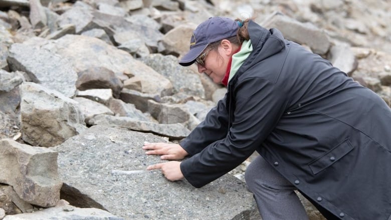 A woman leans on a rock and points at something in or on it.