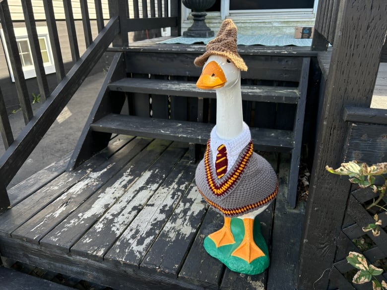 A goose statue wears a grey sweater and a brown witch hat. It is resting on a black wooden porch.