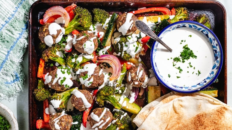 Overhead shot of a sheet pan with meatballs, roasted vegetables, pitas and a small bowl of yogurt tahini sauce.