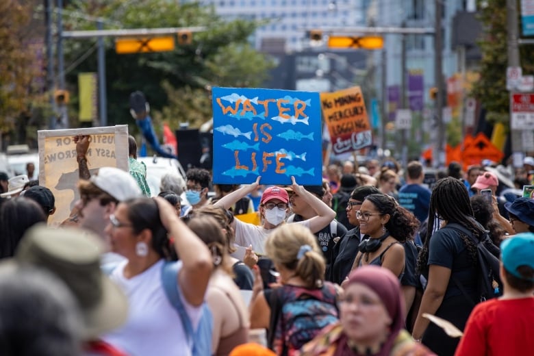 A person is seen in the centre of a large crowd outside, holding a sign that says 