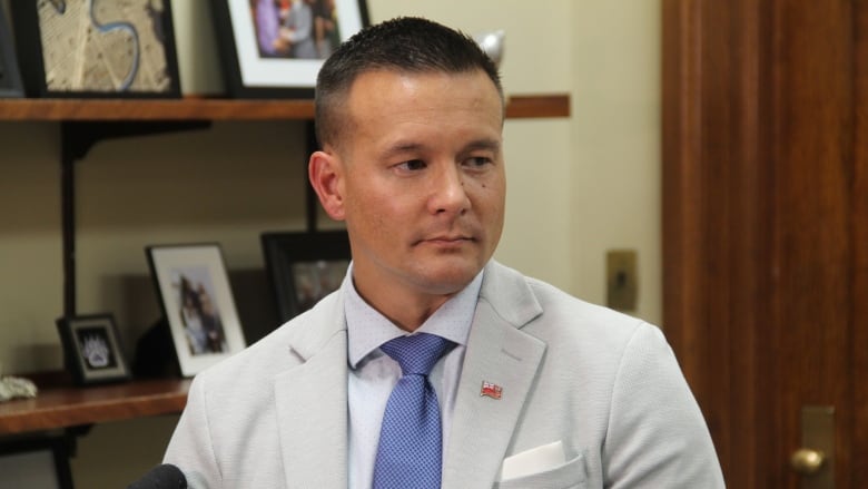 A man in a grey suit and blue tie stands in a room, various framed pictures are beside him.