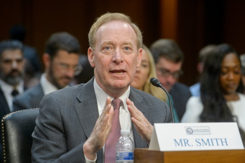 A clean shaven older man in a suit and tie gestures with his hands while sitting a table with a paper placard reading 'Mr. Smith' in front of him.