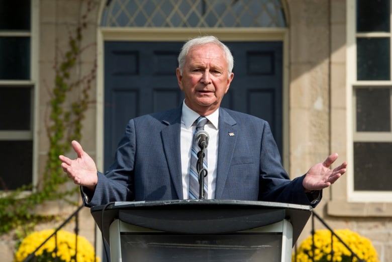 A man in a blue suit stands at a podium outside gesturing with his hands.