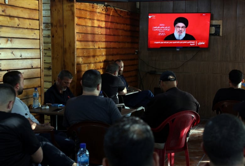 People sitting at a cafe as Nasrallah delivers televised address on the screen.