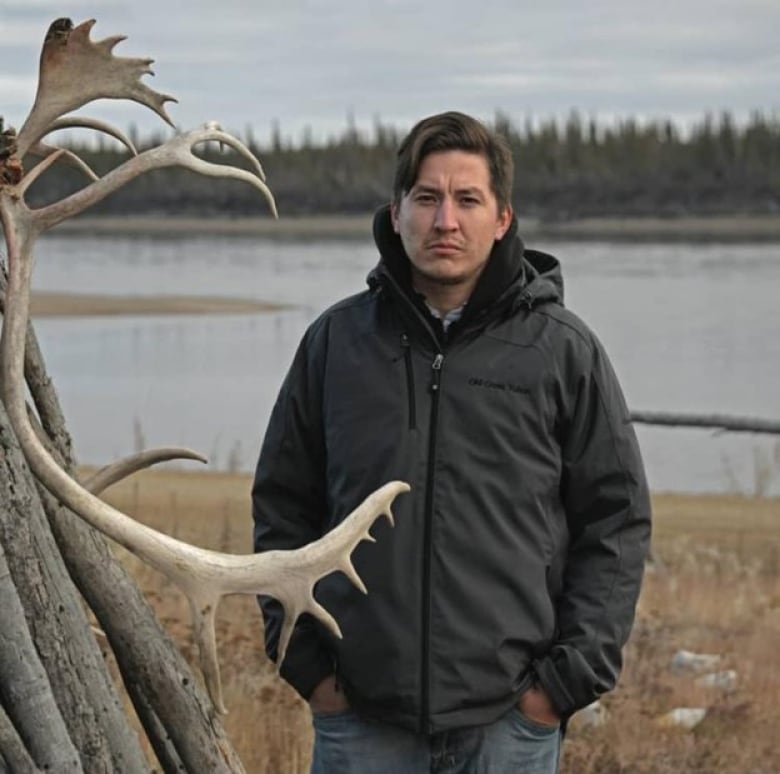 A man in a grey winter jacket stands in front of a river with antlers in the foreground.