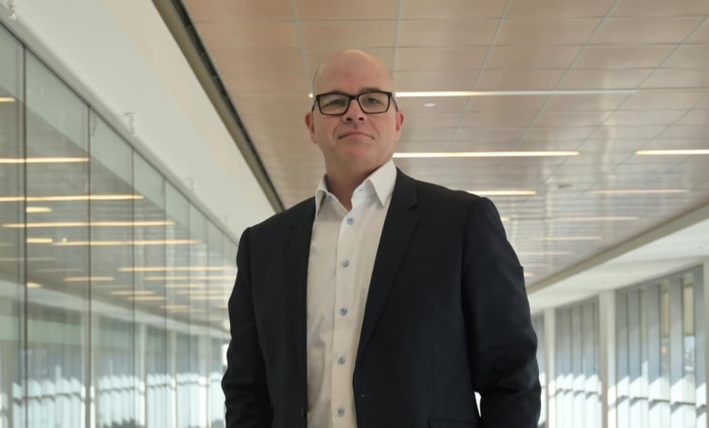 Peter Voros, standing in a long hallway lined with glass windows. He is wearing glasses and a suite. 