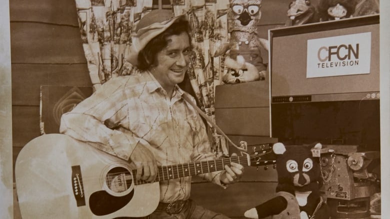 A man wearing a cowboy hat holding a guitar stands next to a puppet.