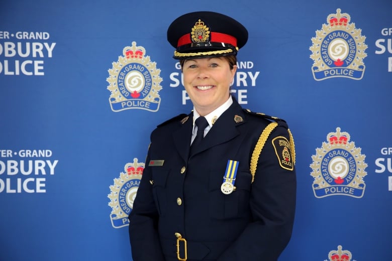 a woman officer in a black police dress uniform smiles while standing in front of a blue background with Greater Sudbury Police logos on it. 