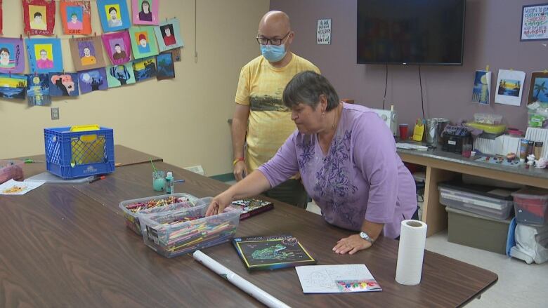 A woman in a purple shirt leans over a table with art supplies.  A man in a yellow shirt and a medical mask stands behind her.