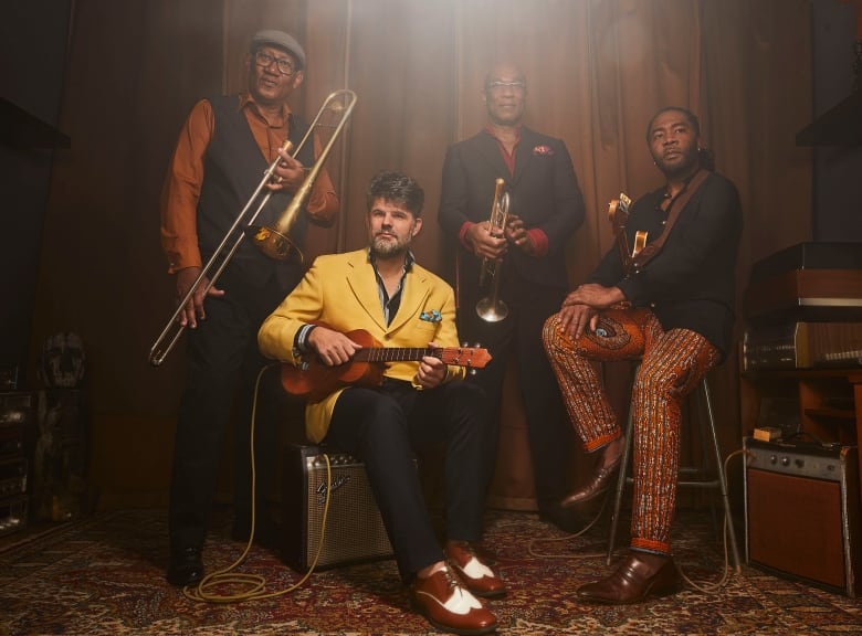 Four men pose for a photo. They are all holding different instruments including a guitar, a trumpet and a trombone.