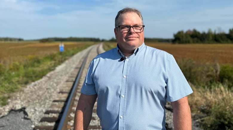 A man in a blue short-sleeved buttoned shirt stands by railroad tracks.