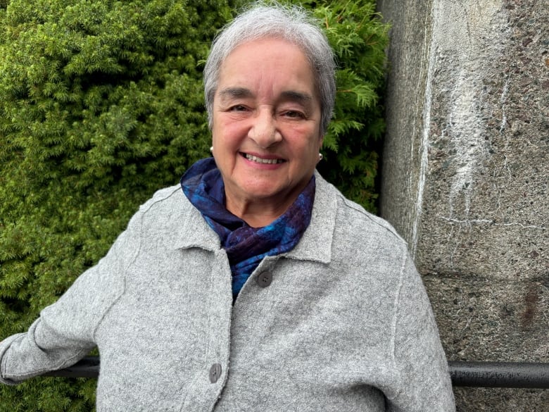 Woman with short grey hair wearing grey button-up sweater and a blue scarf. She's standing against a concrete column and a green bush.