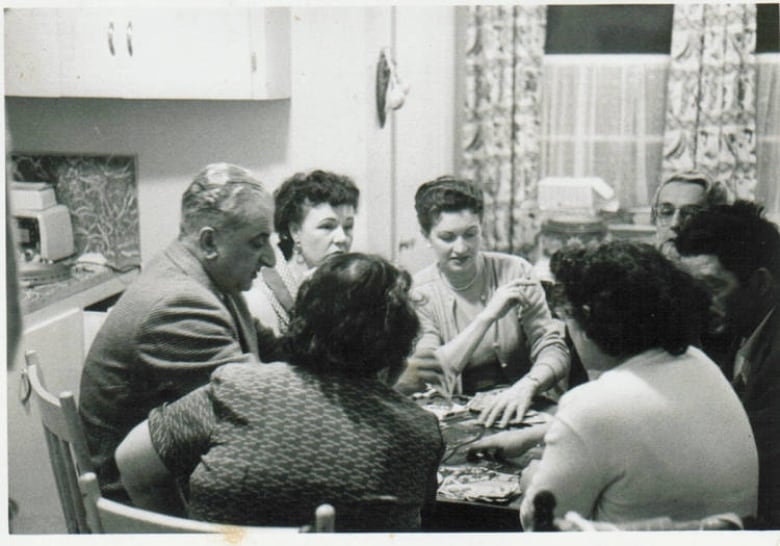 A black and white image of a group gathered around a kitchen table, seated and playing a card game.