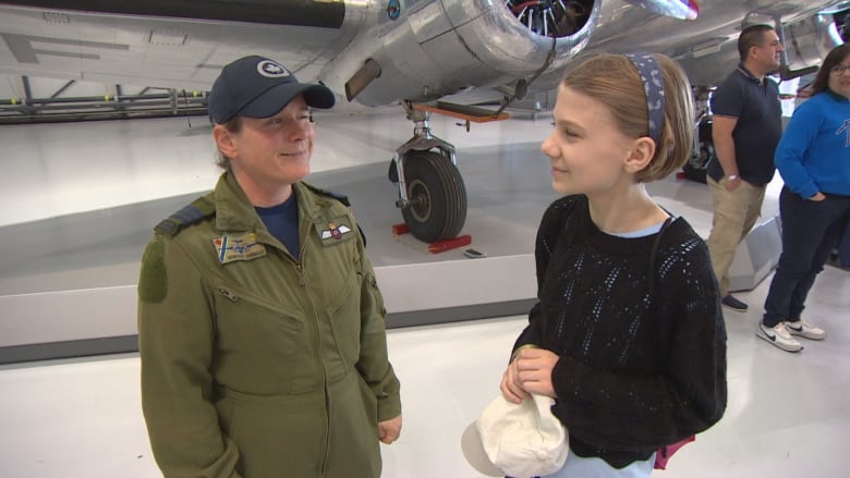 A woman in a green pilot uniform speaks to a young girl with short hair near an airplane. 