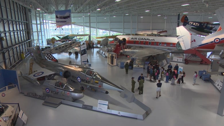 A wide shot of a museum featuring airplanes. A group of people hover near a plane on the right. 