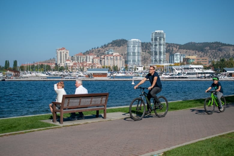 A man and a boy cycle past an older couple sitting on a lakefront bench.