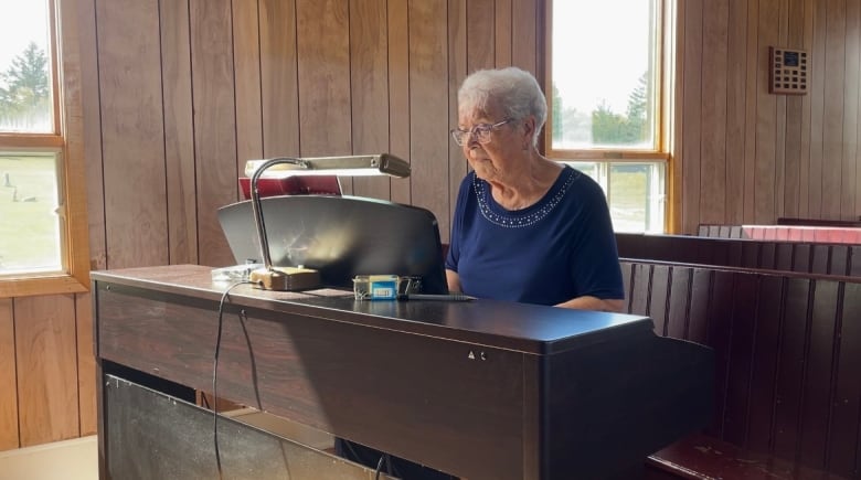 A woman with short white hair and glasses plays the piano in a wooden church