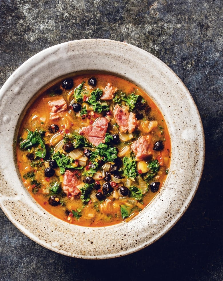 Overhead shot of a shallow bowl of stew with ham, black beans and kale on a dark grey background.