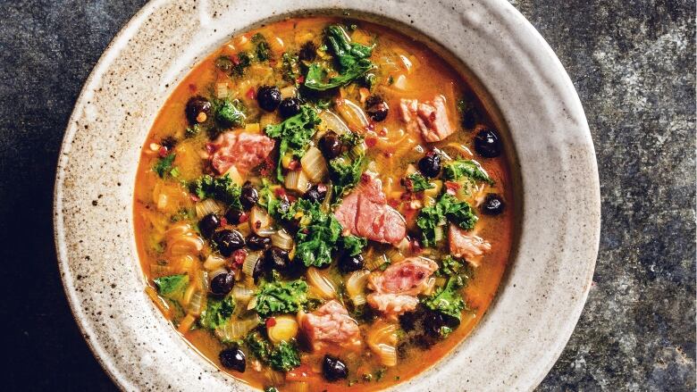 Overhead shot of a shallow bowl of stew with ham, black beans and kale on a dark grey background.