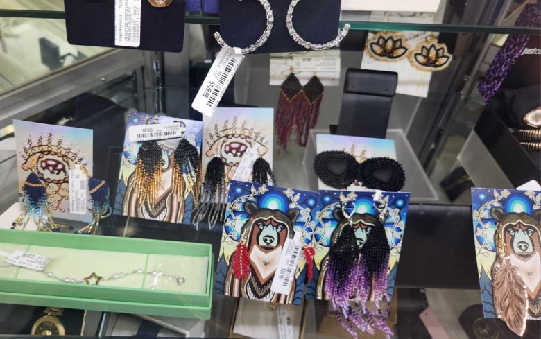 Two rows of beaded earrings, most fringe style in fall coulours such as gray and black, gold, blue and purple on display in a plexiglass case. Also featured, a par of black matte heart earrings surrounded with rows of black beads.