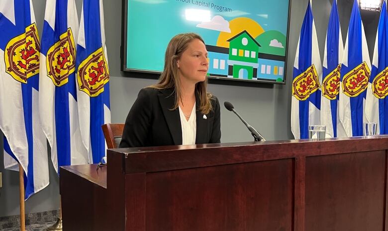 A woman in a blazer sits behind a long wooden podium in front of Nova Scotia flags.