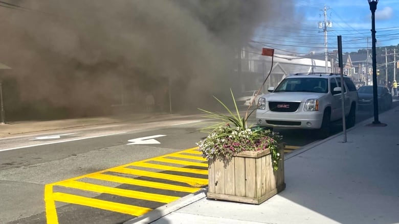 A white car is parked along a street with commercial businesses visible in the background as plumes of black smoke fills the air.