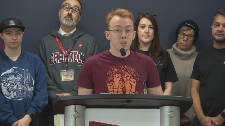 A man in red T-shirt speaks at a podium in a news conference. He has two other people on the left, and three on the right.