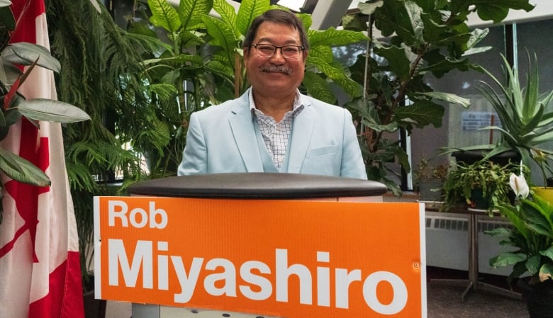 A man wearing a powder blue suit stands behind a podium with an orange sign reading 