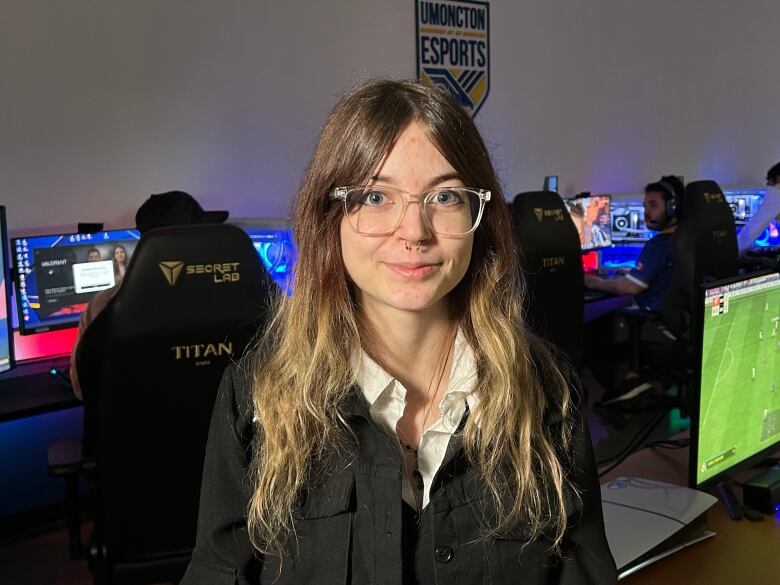 A white lady with blonde hair, wearing glasses, standing in a room with gaming consoles and computers.