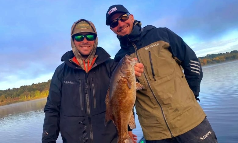 Two men are bundled up and standing ina  boat holding up a fish, smiling into the camera.