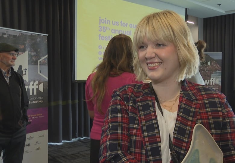 A girl with short blonde hair and a red plaid blazer holds an award that's shaped like a wave.