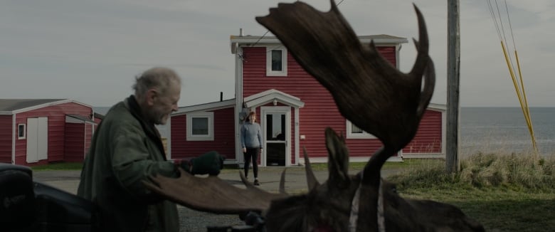 A woman stands near a dead moose. A woman is seen in the background standing in front of a red house.