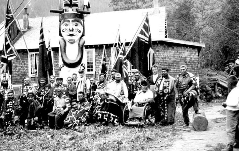 A black-and-white picture of a First Nation band sitting in front of a totem pole, with British flags visible.