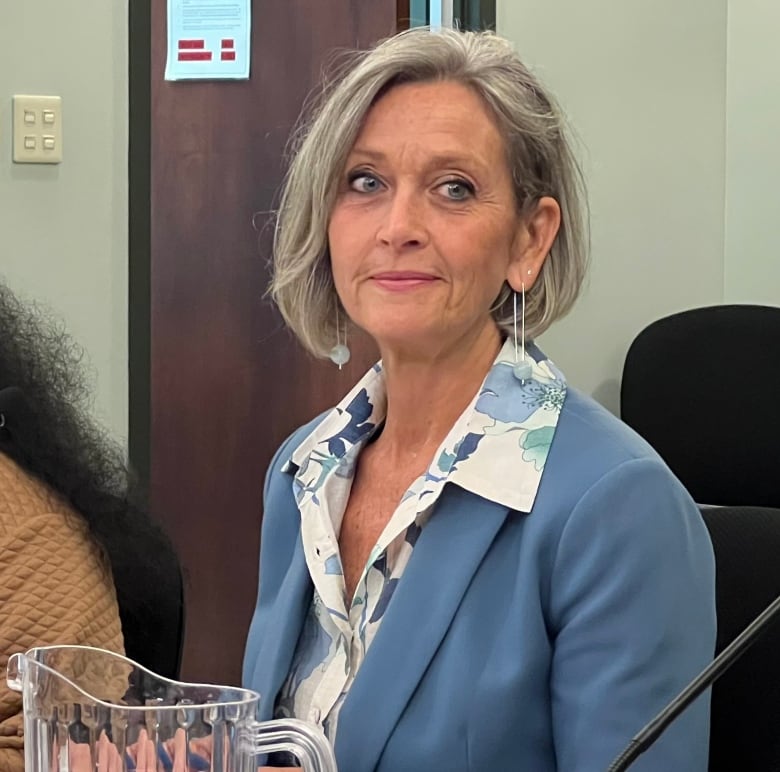 A woman ins a floral shirt and blue blazer sits at a table covered in papers. 