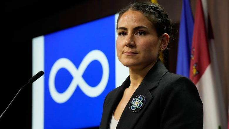 A politician is seen in front of the blue and white Mtis Nation flag.