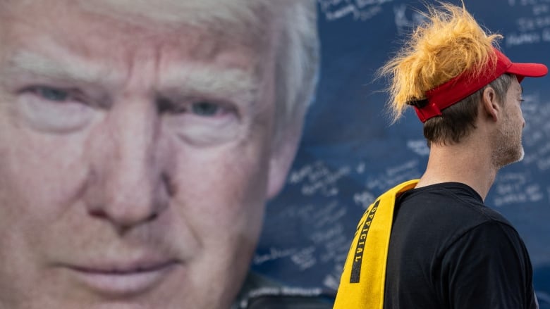 Man in red hat stands in front of big poster with Trump's face on it