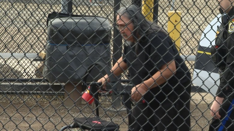 A man with long, grey hair using a walker in a fenced-off police pen