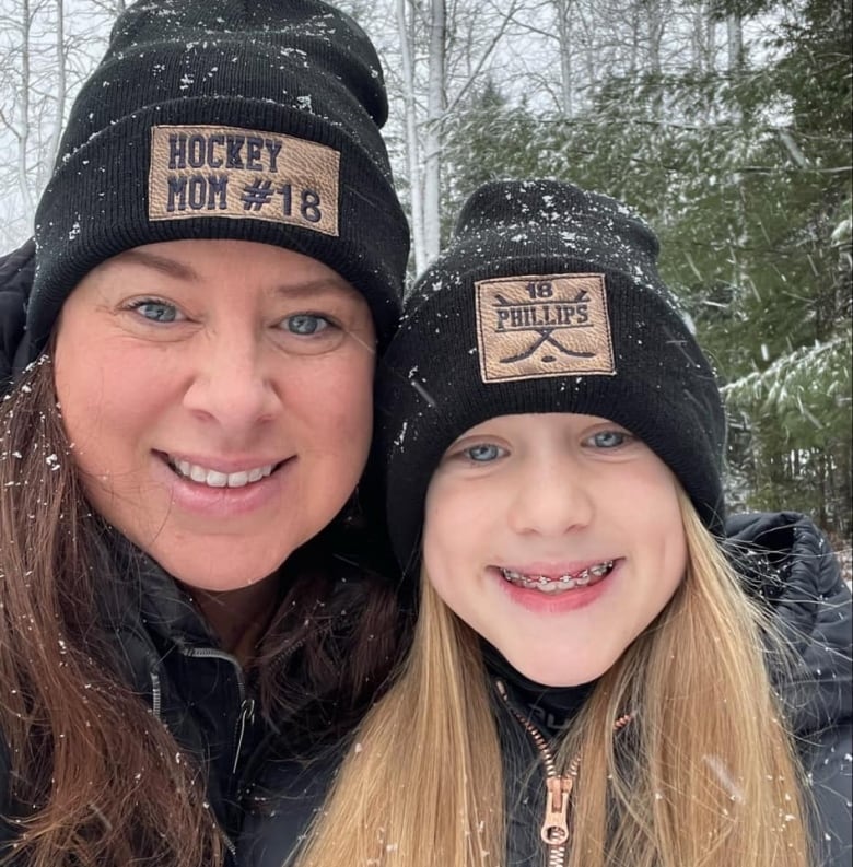 A woman in her 30s with brown hair smiles next to a smiling young girl with blonde hair. Both of them are wearing matching toques. 