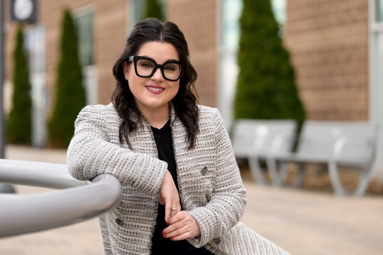 A woman with black hair and black rimmed glasses smiles at the camera, while sitting outside in a dressy jacket. 