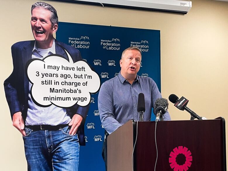 A man speaks into a microphone at a podium, while a larger-than-life cardboard cutout of another man stands behind him.