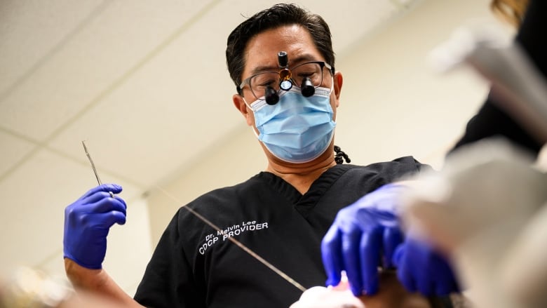 A person wearing surgical loupes, a mask and gloves is holding scissors and sutures. They are looking down at a patient. 