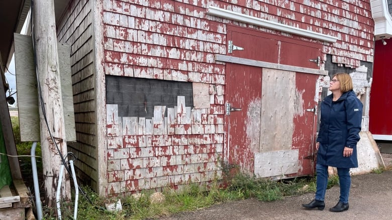 A woman wearing a long blue raincoat looks at a damaged red building on a fishing wharf.